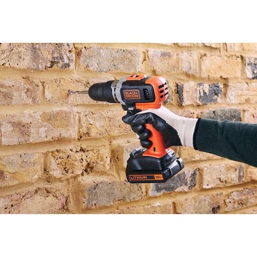 BLACKDECKER BCK21S1S orange cordless drill used on brick wall by gloved hand, 20V lithium battery visible.
