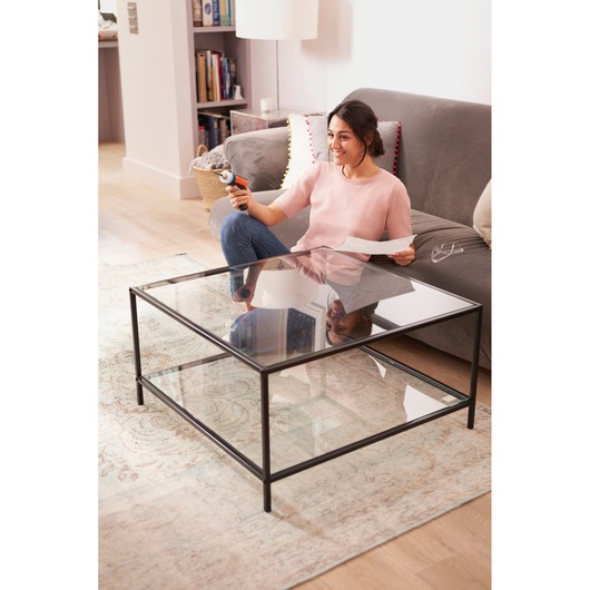 Photo of person using BLACKDECKER BCRTA601I tool while sitting on grey sofa in living room with glass coffee table.