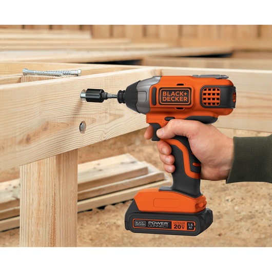 Photo of BLACK+DECKER BDCI20C cordless impact driver in use on wood frames, orange and black design, handheld.