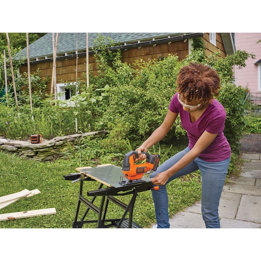 Photo of person using BLACKDECKER cordless jigsaw BCK22S1S to cut wood on a workbench outdoors.