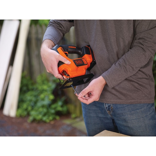 Person holding BLACKDECKER BCK22S1S cordless jigsaw and inserting blade, outdoor workspace in background.