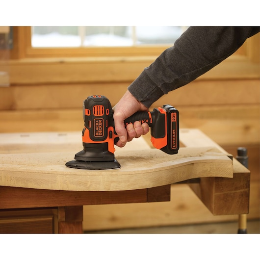 BLACK+DECKER BDCMTS cordless sander in use, sanding a wooden surface in a workshop.