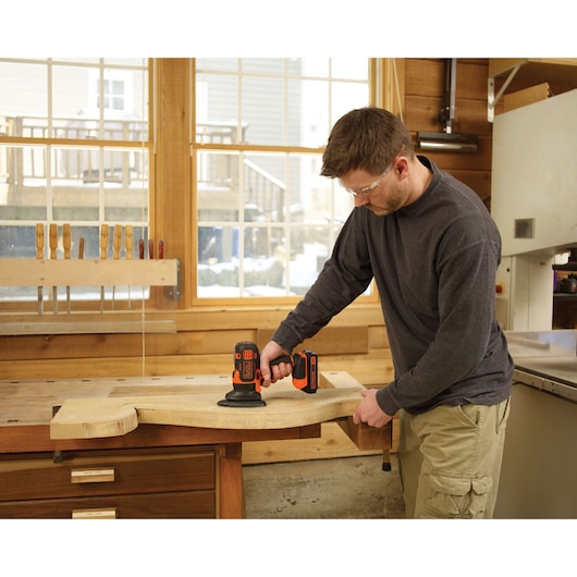 Photo of person using BLACKDECKER BDCMTS cordless sander on wooden board in a workshop.