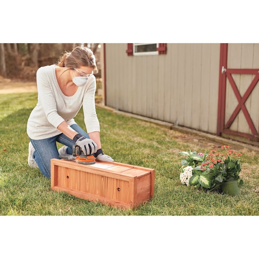 BLACKDECKER BDCRO20C cordless sander used on a wooden box outdoors, flowers and shed in background.