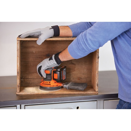 BLACKDECKER BDCRO20C cordless random orbital sander used inside a wooden box, gloved hands visible.