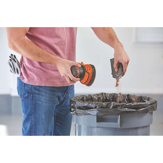 Photo of person emptying dust container from BLACKDECKER BDCRO20C cordless random orbit sander above trash bin.