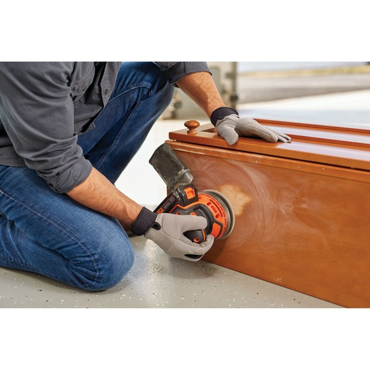 Person sanding wooden chest with BLACKDECKER BDCRO20C cordless random orbital sander, close-up of tool in use.