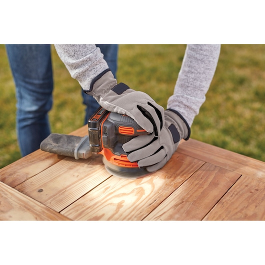 Photo of gloved hands using BLACKDECKER BDCRO20C cordless random orbital sander on wooden table outdoors.