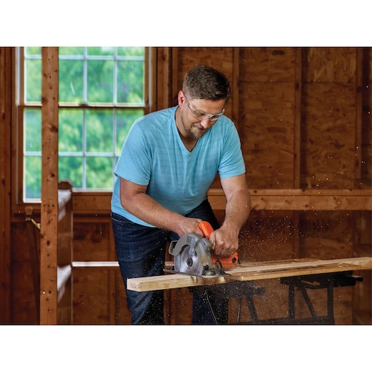 BLACKDECKER BDECS300C circular saw in use, cutting wood on a sawhorse inside a workshop.