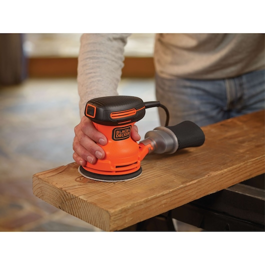 BLACKDECKER BDERO100 orange palm sander in use on wood, close-up angle, power tool photography.