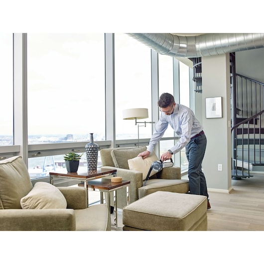 BLACKDECKER BDH2000PL handheld vacuum in use cleaning a beige armchair in a modern living room.
