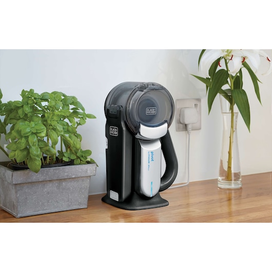 BLACK+DECKER BDH2000PL handheld vacuum on wooden countertop, charging, with a potted plant and vase nearby.