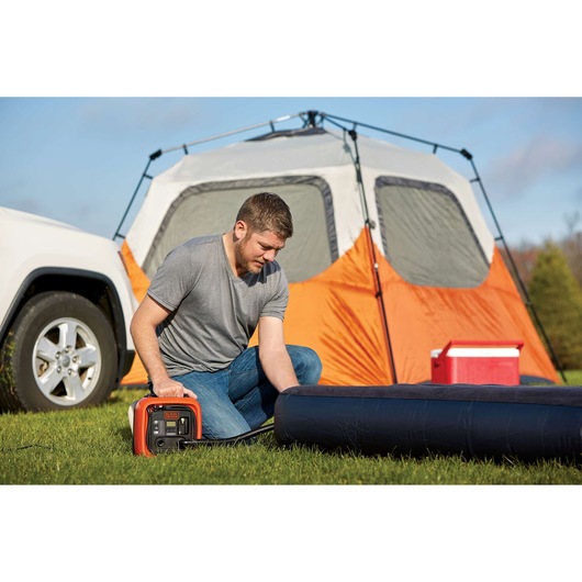 Photo of a person using BLACKDECKER BDINF12C inflator to inflate an air mattress at a campsite with tent and cooler.
