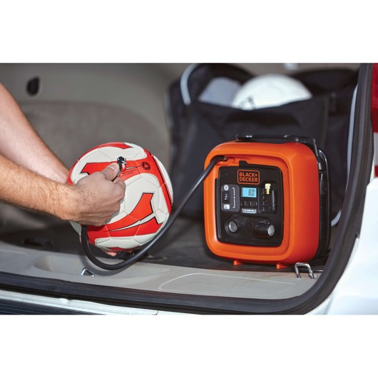 BLACK+DECKER BDINF12C inflator being used to inflate a red and white soccer ball in a car trunk.