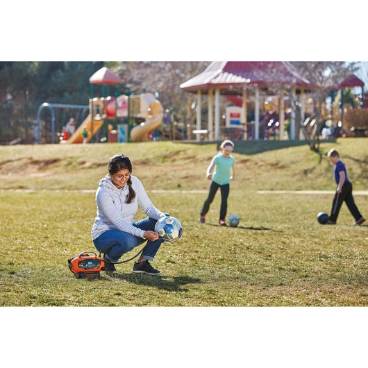 Photo of BLACKDECKER BDINF20 inflator used by a person to inflate a soccer ball on a field, with children playing nearby.