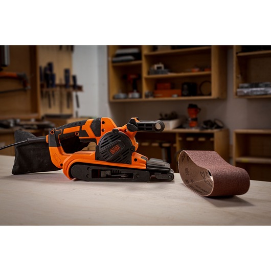 A BLACK+DECKER orange and black belt sander with a dust bag attachment is placed on a workbench. Next to it is a sanding belt. The background shows shelves and tools in a workshop.