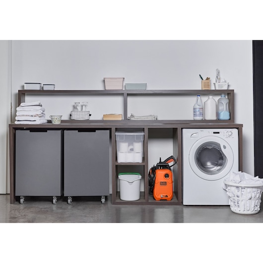 A laundry room setup featuring a washing machine, two gray storage bins on wheels, shelves with laundry supplies and towels, a white basket with clothes, and an orange BLACK+DECKER pressure washer (model BEPW1600) placed on the lower shelf.