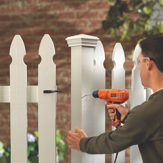 Person using BLACKDECKER DR260C corded drill on white wooden fence outdoors.