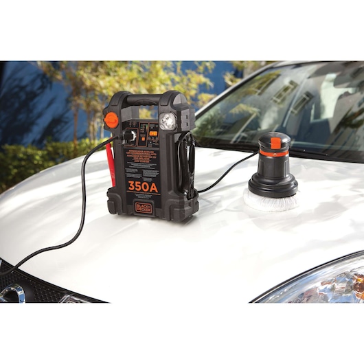A BLACK+DECKER portable jump starter (model JS350CC, 350A) with cables and a buffer/polisher attachment is placed on the hood of a white car, outdoors.