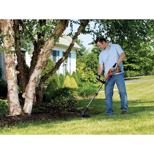 Person using BLACKDECKER LST136 cordless string trimmer in a garden next to trees and lawn.