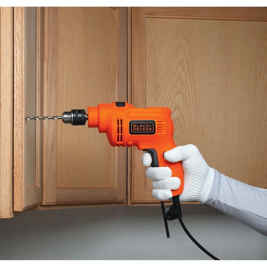 Photo of BLACK+DECKER TP550 orange corded drill in use on a wooden cabinet, held by a gloved hand.